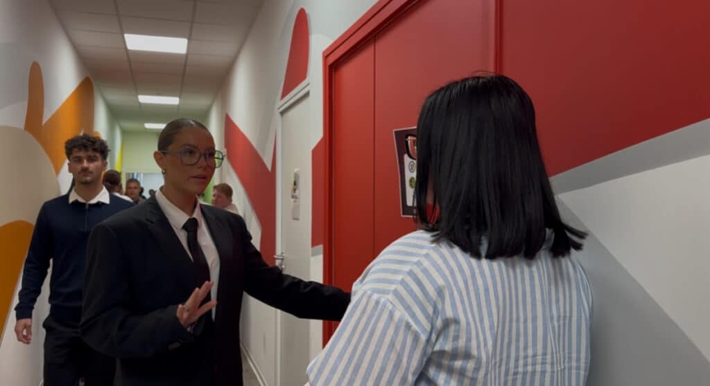 Un couloir où une candidate et un membre du staff sont en train de discuter. Le membre du staff rassure la candidate avant son passage.