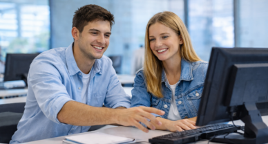 Deux étudiants échangent autour d’un ordinateur dans un environnement de bureau lumineux, concentrés et souriants.